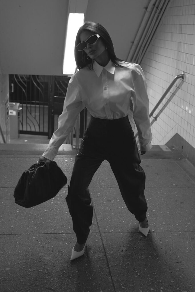 Victoria Barbara street style in New York City subway wearing white Bottega Veneta blouse top, Khaite black pants, black Bottega Veneta pouch handbag, white Balenciaga heeled shoes, & Loewe sunglasses during New York Fashion Week 2021