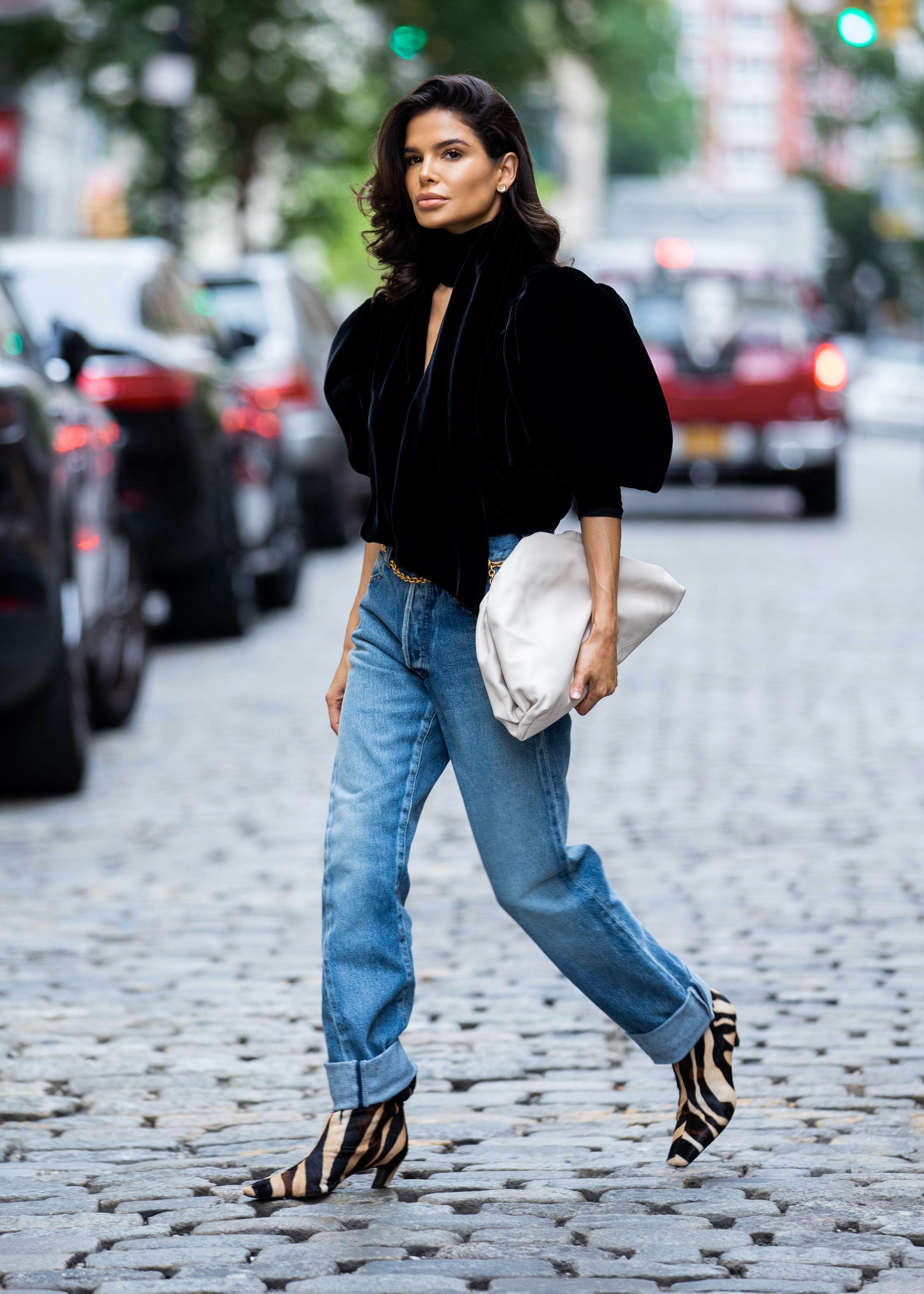 Victoria Barbara walking along a cobblestone street during New York Fashion Week wearing a black KHAITE velvet top, Balenciaga jeans, Chanel chain belt, Bottega Veneta clutch and KHAITE zebra-print boots.