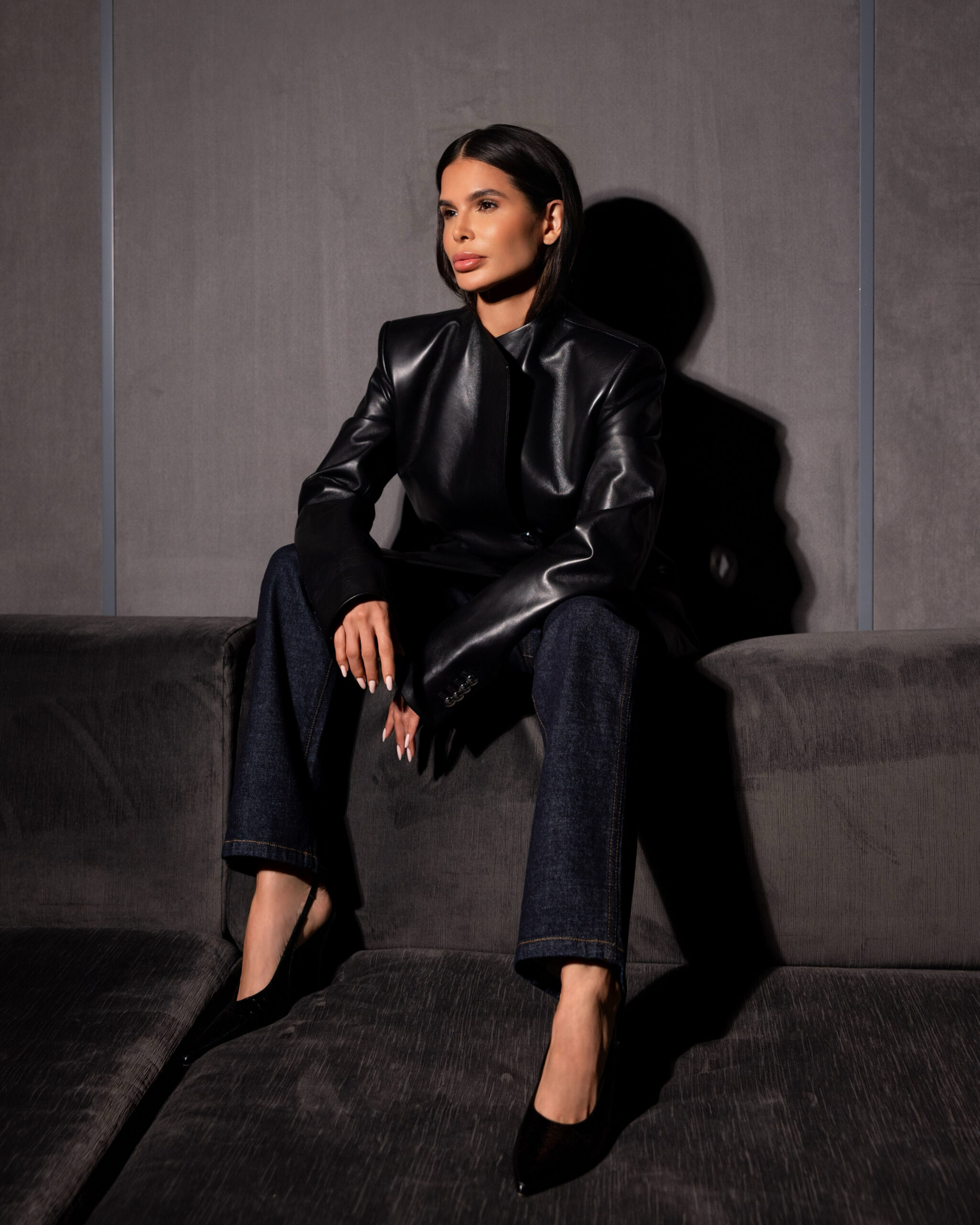 Khaite editorial look featuring structured black leather blazer and dark denim, seated pose with architectural lighting and shadow