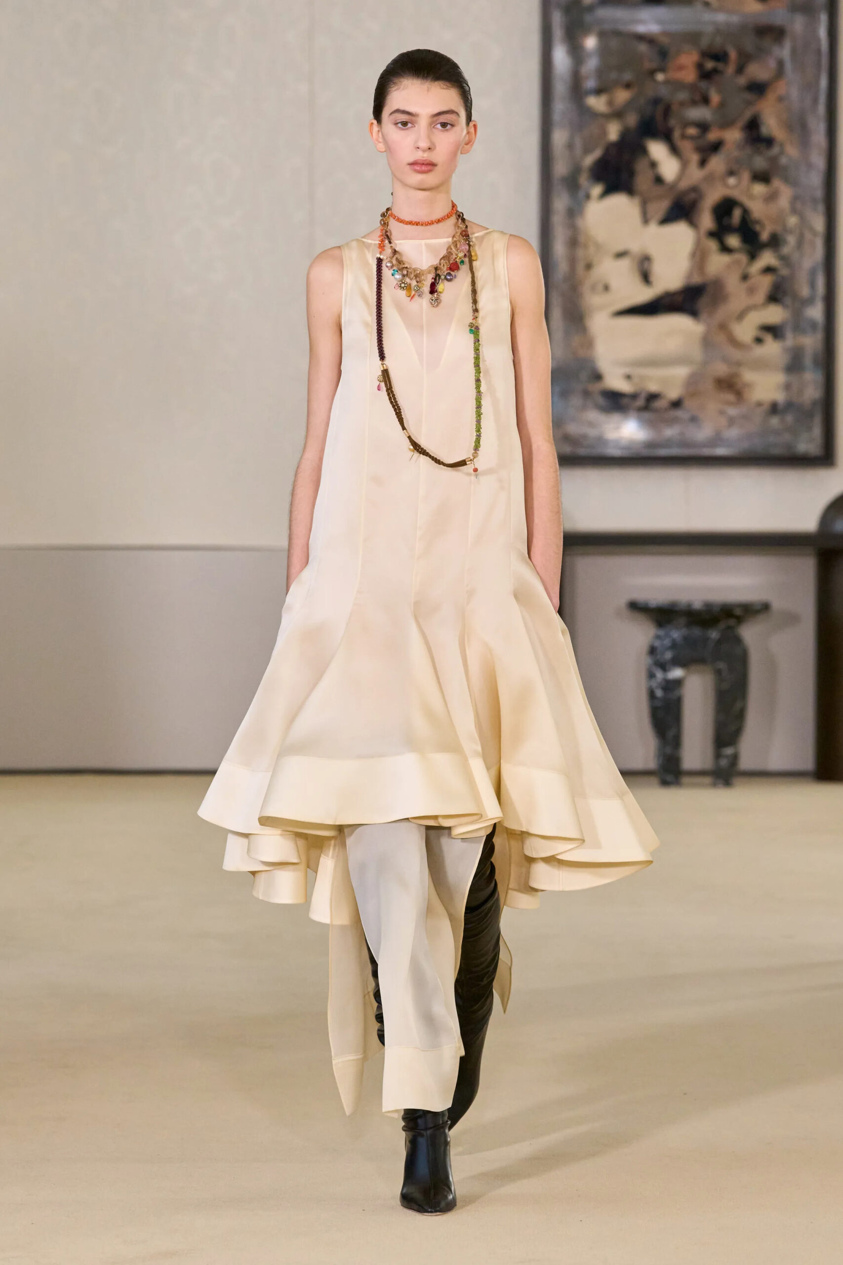Altuzarra Fall Winter 2026 runway look featuring an asymmetrical ivory draped dress layered over trousers with black leather boots and layered multicolored necklaces at New York Fashion Week