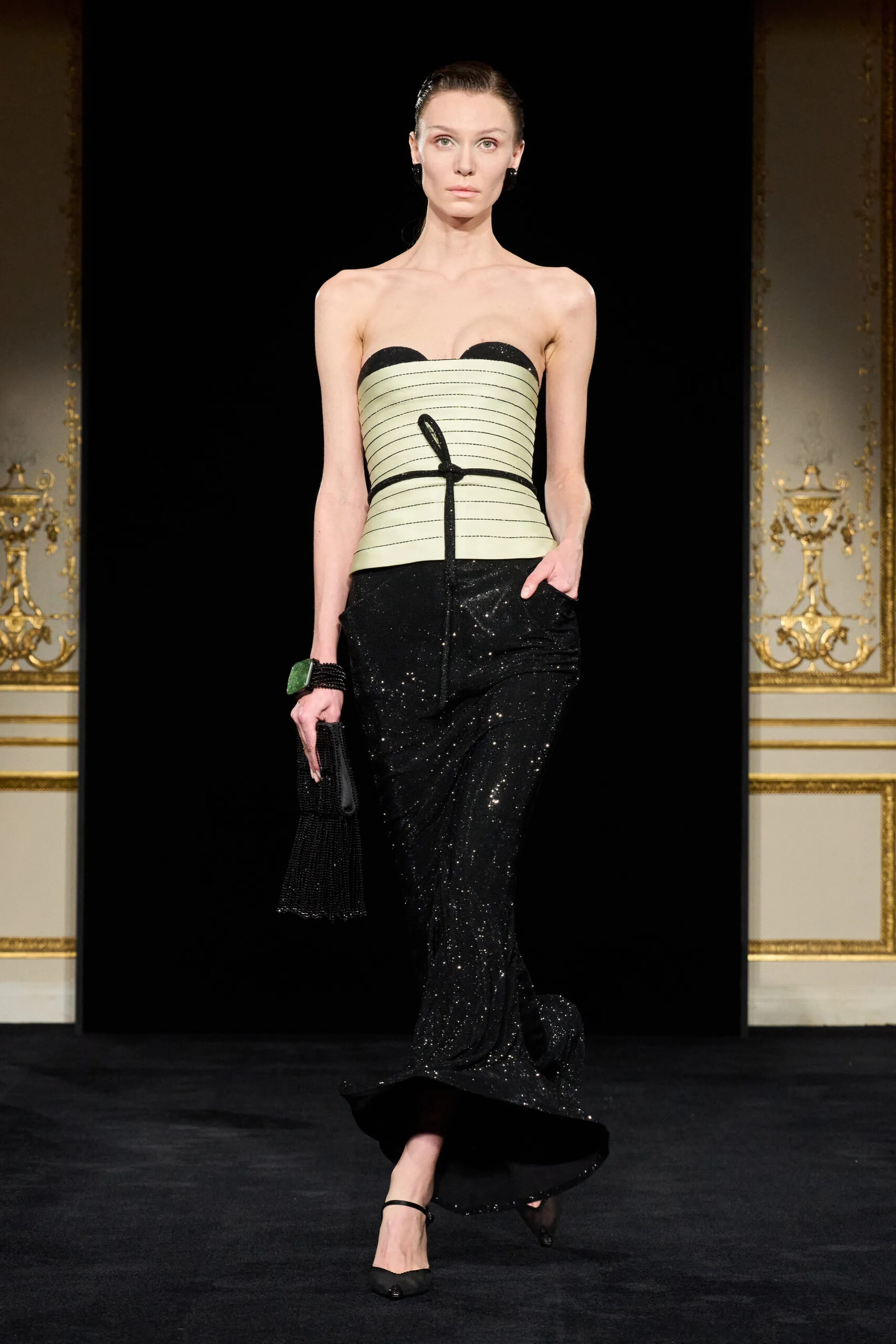 Model walking in Armani Privé Spring 2026 Couture wearing a pale mint structured corset top with black sequined column skirt, black accessories, and pointed heels against a dark runway backdrop