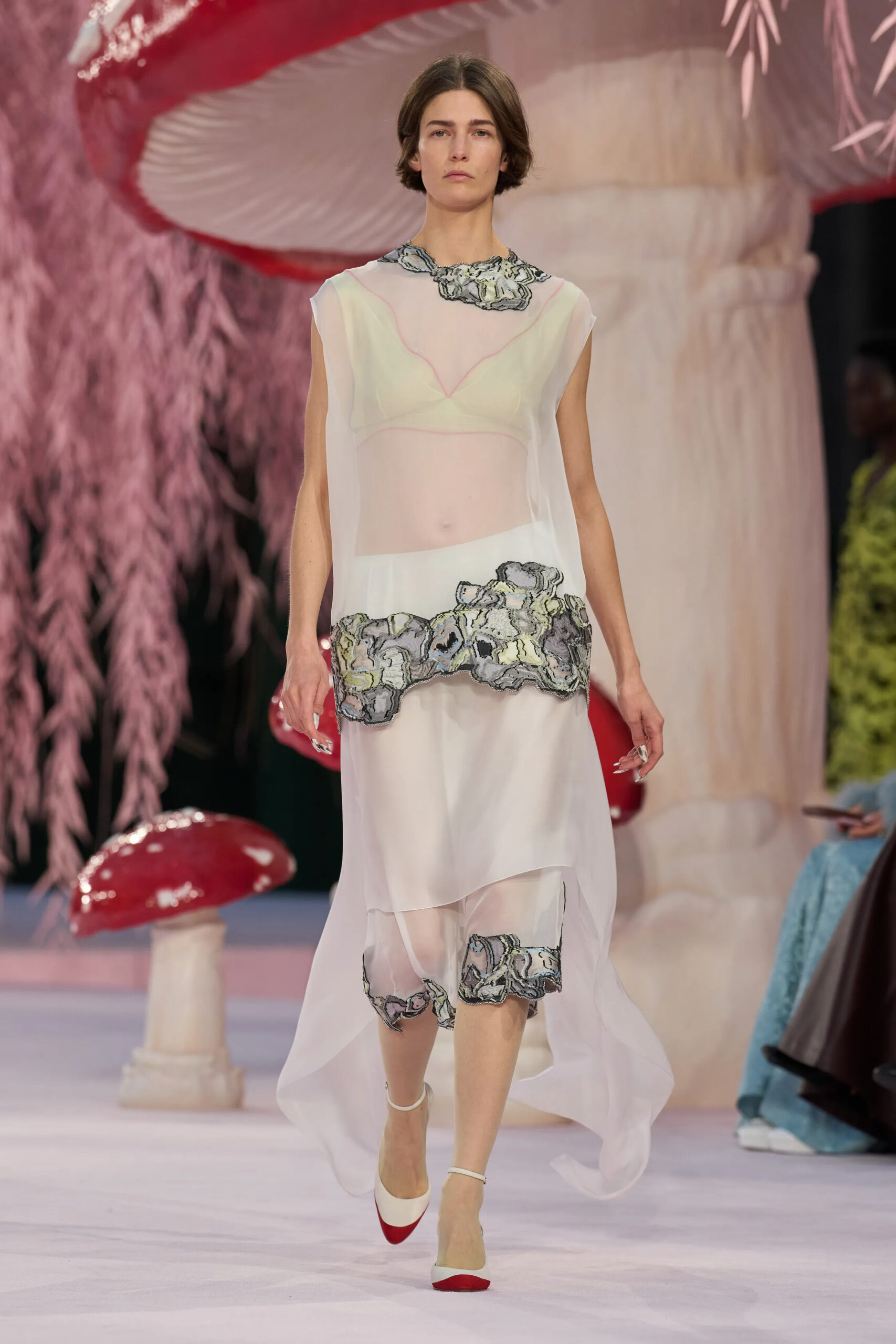 Model wearing a sheer white Chanel couture dress with abstract embroidered detailing on the runway