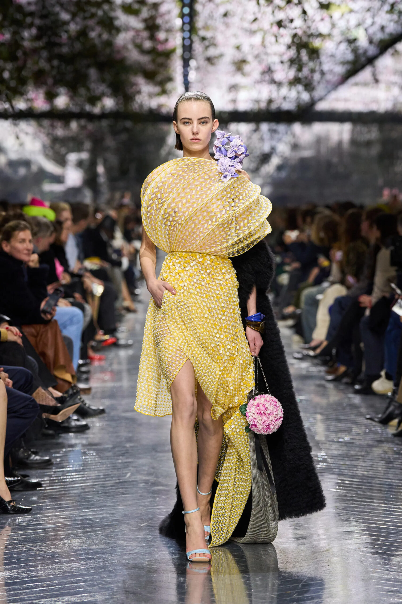 Model walking in yellow Dior couture dress with sculptural pleated bodice and lavender floral accent