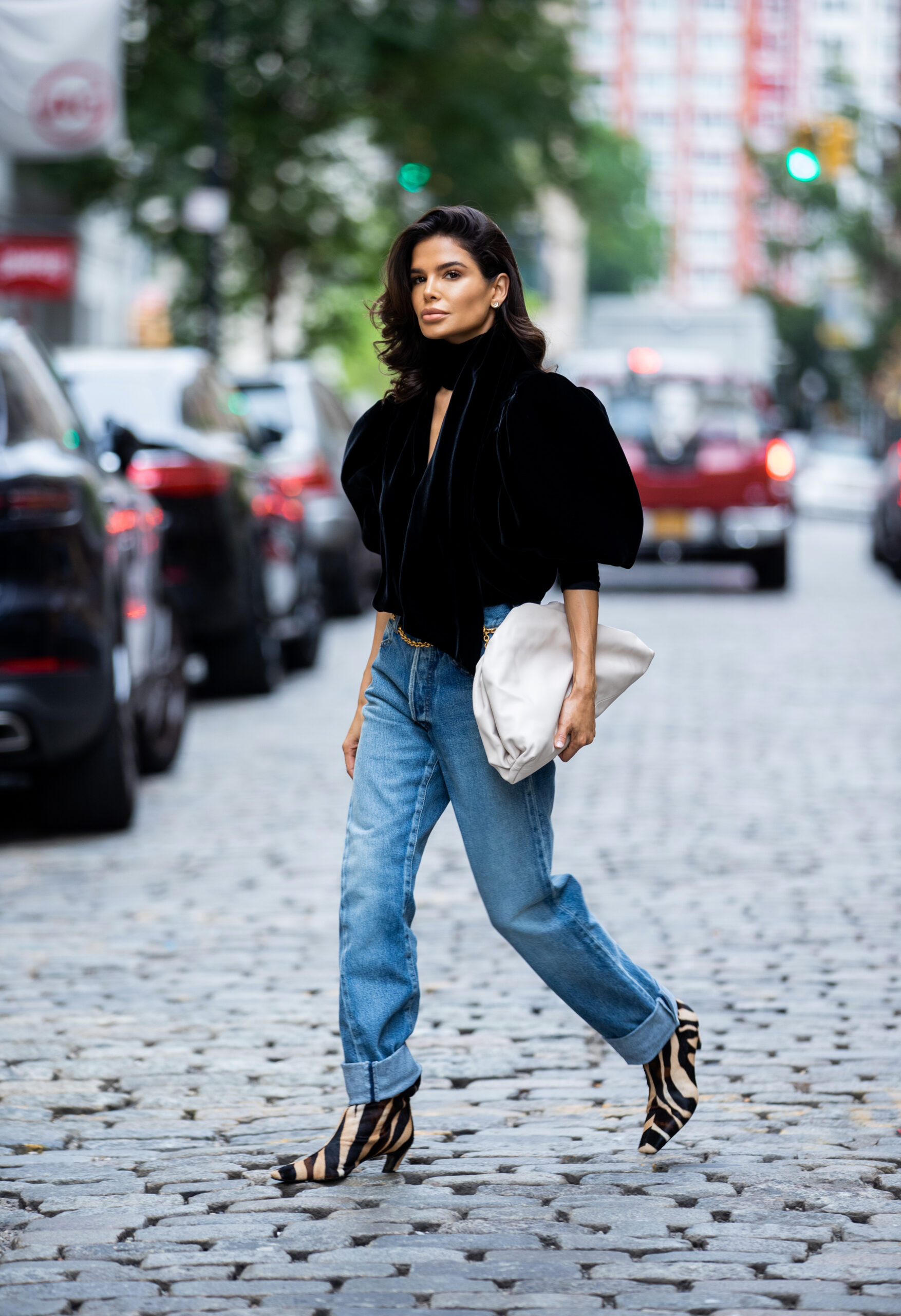 Victoria Barbara walking along a cobblestone street during New York Fashion Week wearing a black KHAITE velvet top, Balenciaga jeans, Chanel chain belt, Bottega Veneta clutch and KHAITE zebra-print boots.