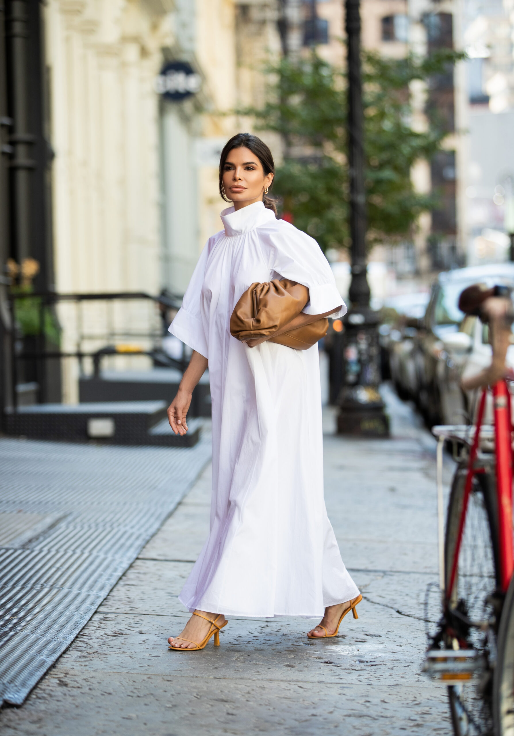 Victoria Barbara walking through New York wearing a white The Row dress with a structured high collar, styled with a brown Bottega Veneta clutch and The Row sandals.
