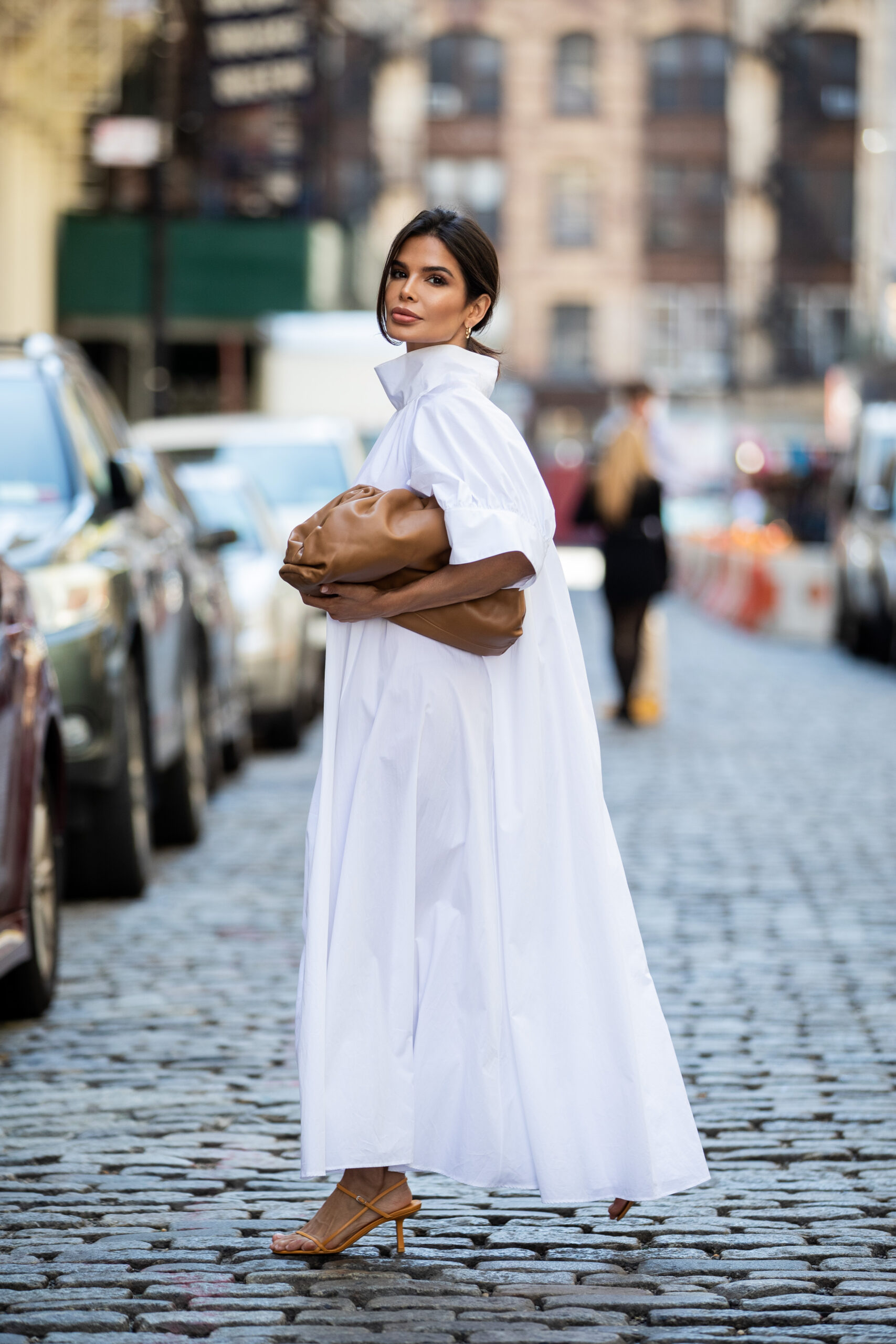 Victoria Barbara photographed on a cobblestone New York street wearing a white The Row dress with a Bottega Veneta clutch tucked under her arm.