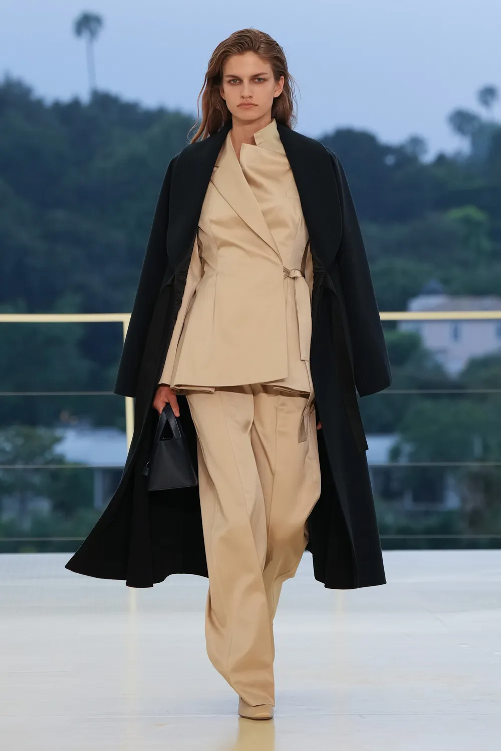 Model wearing an ivory wrap jacket and wide-leg trousers layered beneath a black overcoat, carrying a structured handbag at the Hermès Fall-Winter 2026 show.