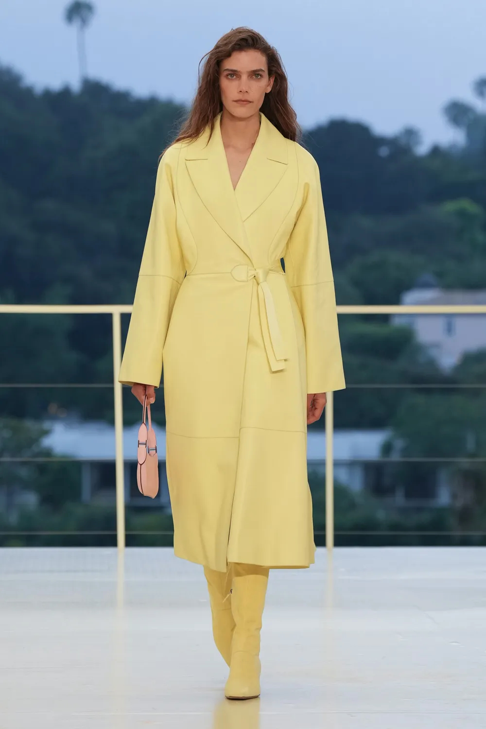 Model walking the runway in a butter yellow belted coat paired with matching knee-high boots and a pale pink handbag at the Hermès Fall-Winter 2026 show.