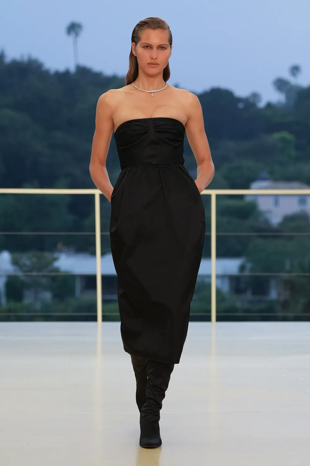 Model wearing a black strapless dress with a diamond necklace and knee-high black boots on the Hermès Fall-Winter 2026 runway.
