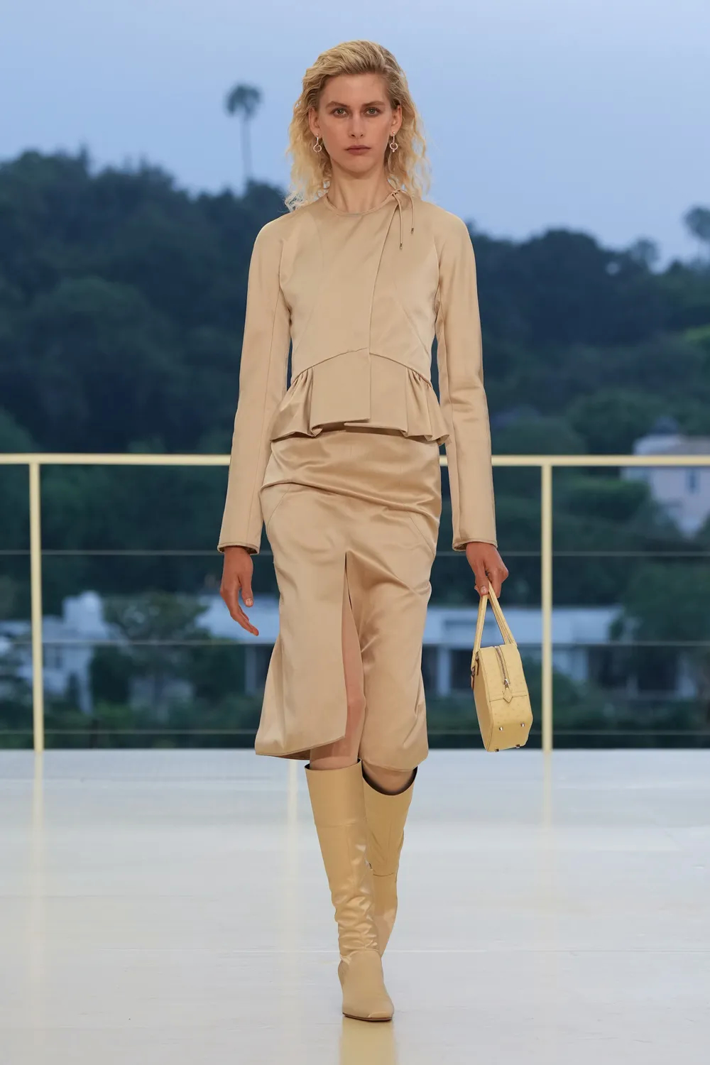 Model wearing a champagne-colored dress with draped detailing, matching handbag, and cream leather boots at the Hermès Fall-Winter 2026 show.