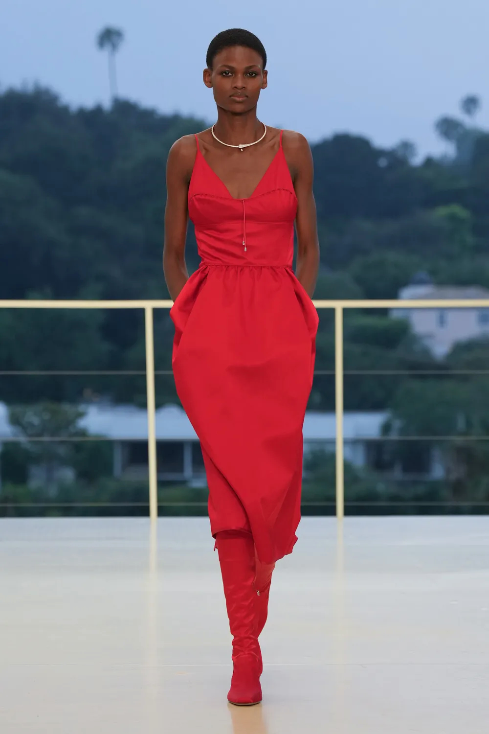 Model walking the runway in a bright red sleeveless dress paired with matching knee-high boots and delicate jewelry at the Hermès Fall-Winter 2026 show.