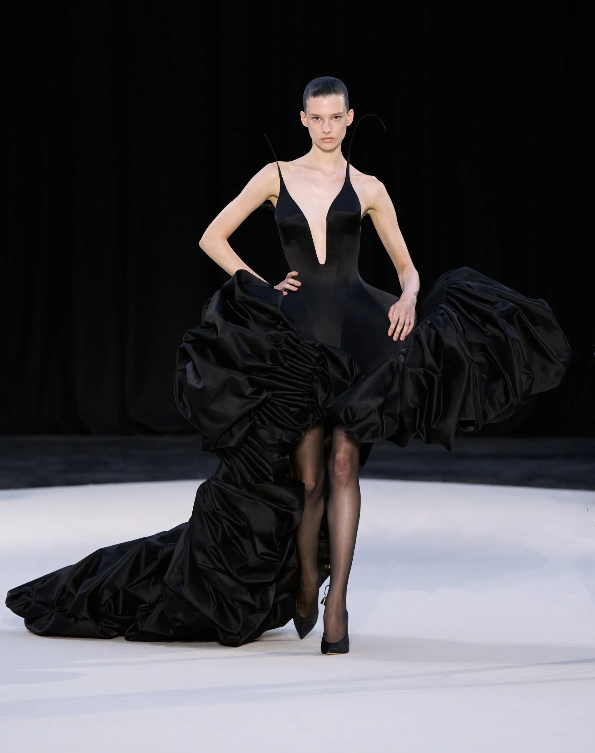 Model wearing a black Ashi Studio Fall/Winter 2026 Couture gown featuring a plunging neckline, sculptural oversized satin side draping, sheer tights, and a sweeping train presented on the Paris Haute Couture runway