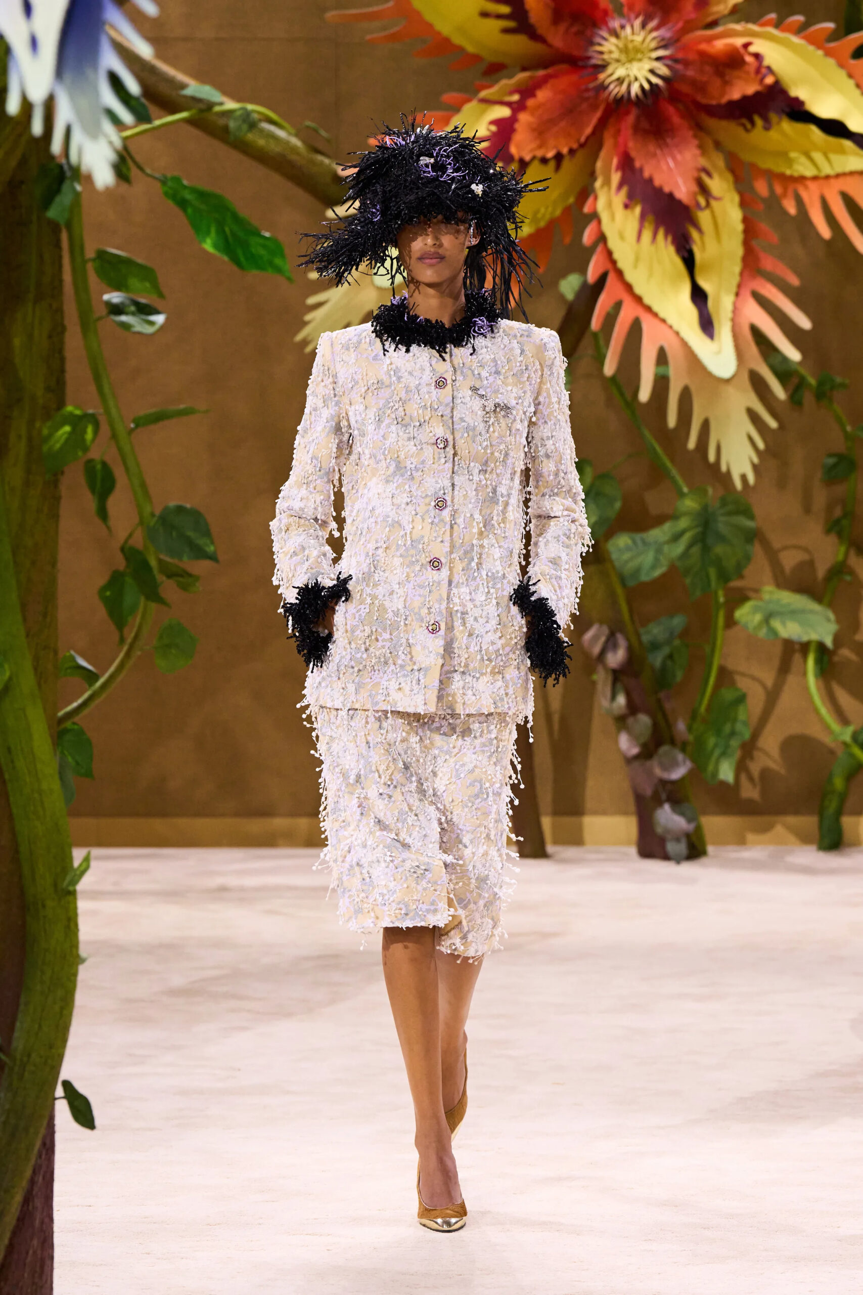 Model wearing a white textured Chanel couture skirt suit with black feathered trim and an oversized feathered hat