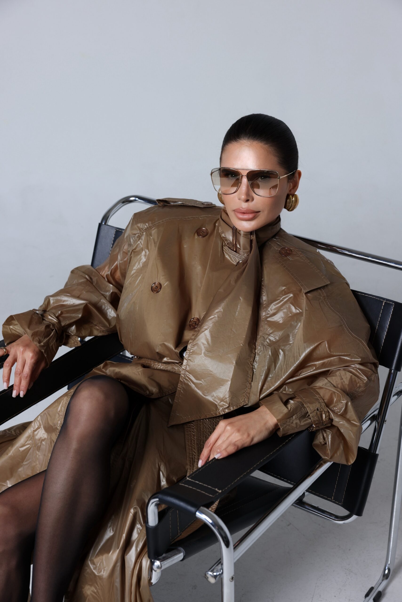 Victoria Barbara wearing Saint Laurent in a sculptural editorial portrait featuring an oversized trench coat, sheer tights, pointed pumps, and modernist seating